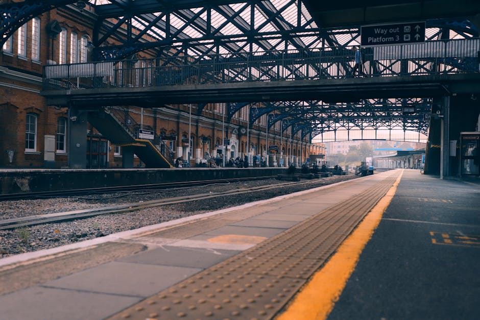 The image depicts a spacious train station platform view with a metal and glass arched roof structure overhead, supported by blue-painted steel beams. On the left, the historic station building features red brick walls, large rectangular windows, and architectural details typical of traditional UK railway stations. The platform surface consists of concrete with tactile paving strips in yellow along its edge for safety, guiding passengers toward the train. In the background, train tracks run parallel along the platform, extending into the distance, with a few small trains and station signage visible. Above the platform, illuminated directional signs are mounted, indicating platform information and exit routes. The scene is lit by natural daylight filtering through the glass roof, creating an even, diffuse lighting atmosphere. The overall environment suggests a well-maintained, busy transportation hub designed for passenger comfort, with nearby urban buildings and infrastructure in the distance. This detailed view aligns with a focus on infrastructure that could support various aspects of passenger and freight logistics, including alternative or private waste handling linked to station operations or maintenance areas, relevant to waste collection services such as those by Waste Collection Bermondsey.