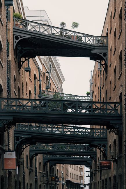 The image shows a narrow city street flanked by tall, multi-storey brick buildings with a series of black-painted iron pedestrian bridges connecting the upper floors. The bridges have decorative railings and structural supports visible beneath, with some small potted plants placed along their edges. The buildings feature numerous evenly spaced rectangular windows, some with black window frames, and the brickwork has a reddish-brown tone with slight weathering. The sky above is overcast, providing diffuse natural lighting that highlights the textures of the materials. The scene evokes an urban environment where private or alternative ways of connecting different building levels are employed, reflecting a design that may facilitate access or aesthetic appeal. The street below appears quiet, with no visible pedestrians or vehicles, emphasizing the architectural detail of the bridges and surrounding facades, which are typical features in historic or industrial-inspired cityscapes related to waste collection or site management contexts by companies like Waste Collection Bermondsey, operating within the premise of rubbish removal and property clearance services.