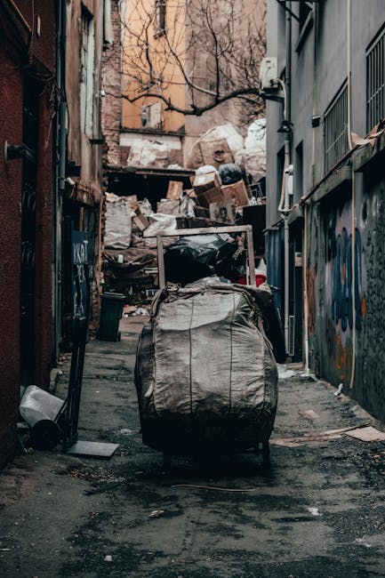 A large black and gray wheeled trash bag with a crinkled surface sits in the center of a narrow, grimy alleyway with a concrete floor that shows signs of staining and debris. Surrounding the bag, various discarded items such as a black plastic container and miscellaneous debris are visible on the ground. In the background, a pile of mixed waste including cardboard boxes, plastic bags, and other rubbish is stacked against an unfinished wooden pallet or makeshift barrier. The alley is flanked by tall, weathered buildings with graffiti on the right wall and exposed piping and fixtures on the left. Bare tree branches extend overhead, and a muted, overcast sky is visible above, creating an overall dim and gritty urban atmosphere. The scene appears to capture an outdoor waste collection or clearance area, consistent with private rubbish removal services like those provided by Waste Collection Bermondsey, operating within a typical urban environment dedicated to rubbish disposal and collection.