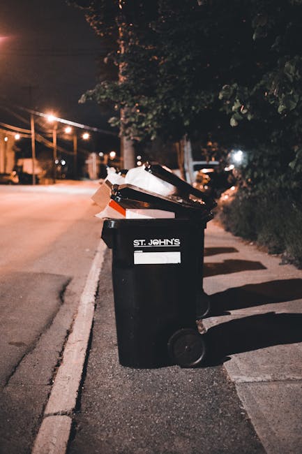 A black wheeled rubbish bin with the label 'St. John's' positioned on a sidewalk beside a curb in an urban area during nighttime. The bin is filled with mixed waste, including paper, cardboard, and plastic items, some of which are overflowing and leaning outward. The surface of the bin appears slightly textured with a matte finish. Behind the bin, there are shadows cast by nearby trees and streetlights, creating a dimly lit environment. In the background, a quiet residential street is visible, illuminated by warm street lighting, with a row of trees and parked vehicles along the roadside. The concrete sidewalk has minor cracks and a white-painted line marking the edge of the roadway. This scene exemplifies an independent waste collection point, potentially managed by private waste services like Waste Collection Bermondsey, reflecting typical rubbish disposal practices in urban settings.