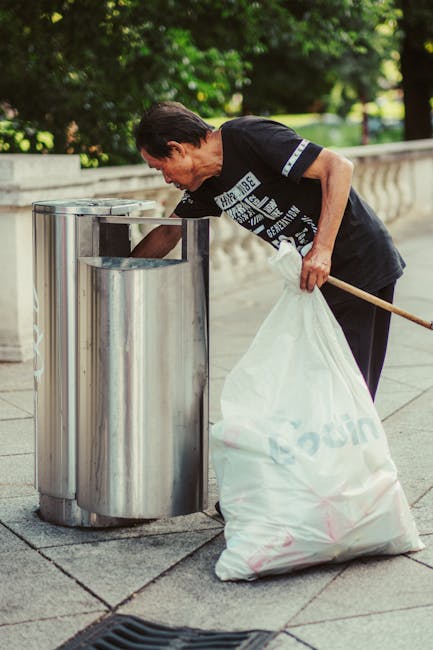 A large black and gray wheeled trash bag with a crinkled surface sits in the center of a narrow, grimy alleyway with a concrete floor that shows signs of staining and debris. Surrounding the bag, various discarded items such as a black plastic container and miscellaneous debris are visible on the ground. In the background, a pile of mixed waste including cardboard boxes, plastic bags, and other rubbish is stacked against an unfinished wooden pallet or makeshift barrier. The alley is flanked by tall, weathered buildings with graffiti on the right wall and exposed piping and fixtures on the left. Bare tree branches extend overhead, and a muted, overcast sky is visible above, creating an overall dim and gritty urban atmosphere. The scene appears to capture an outdoor waste collection or clearance area, consistent with private rubbish removal services like those provided by Waste Collection Bermondsey, operating within a typical urban environment dedicated to rubbish disposal and collection.