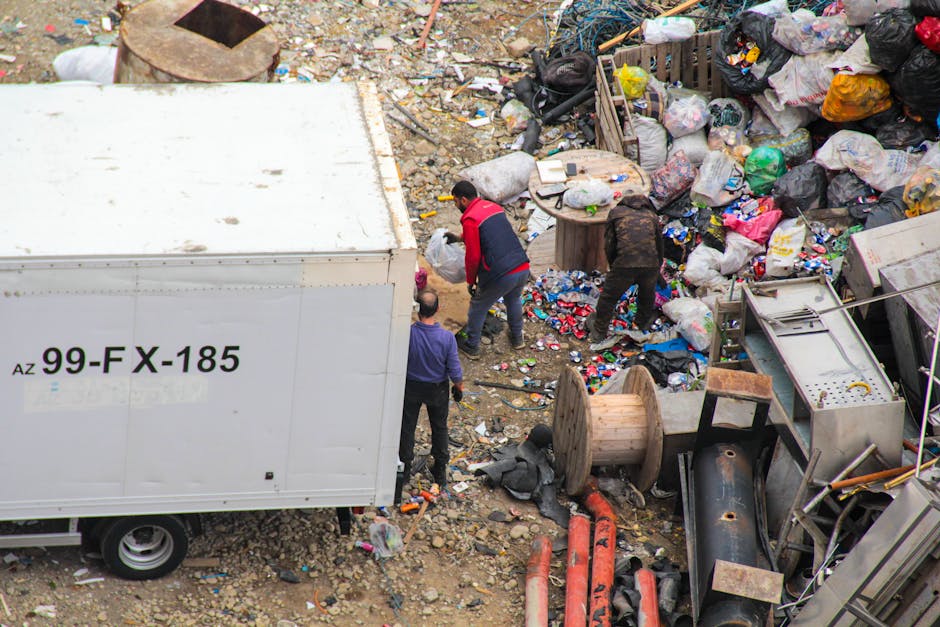 The image shows an outdoor scene with three individuals sorting through a large pile of mixed waste and rubbish. The waste consists of plastic bags, cardboard, metal, and various miscellaneous debris scattered across the ground. One person, wearing a red and black jacket, is actively engaged in handling or inspecting the waste, while another, dressed in dark clothing, appears to be rummaging through the rubbish. A third individual, dressed in a purple garment, is standing nearby observing or assisting. Adjacent to the waste pile, there is a large white shipping container marked with the code 'AZ 99-FX-185,' which is positioned close to the sorting activity. The surrounding environment includes an uneven, rocky surface with small stones and discarded materials, indicating an area set aside for waste collection or clearance. There are also visible large industrial or construction objects, such as spools, pipes, and partially tipped or overturned equipment, contributing to the appearance of a site dedicated to on-site rubbish clearance or waste handling, consistent with services provided by waste collection specialists like Waste Collection Bermondsey. The overall scene captures a moment of manual waste sorting, aligned with private or alternative rubbish removal methods in an outdoor industrial or construction setting.