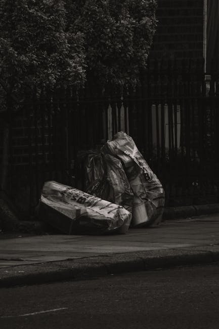 Two large black plastic bags filled with waste materials are positioned on the edge of a paved sidewalk, leaning against a black wooden fence with vertical slats. The bags appear crumpled and contain a mixture of recyclable and non-recyclable rubbish, including flattened cardboard boxes and plastic packaging. In the background, there are dense leafy bushes and trees with dark foliage, partially obscuring a structure behind the fence. The scene is lit by natural daylight, with shadows cast on the ground and objects, indicating a clear day. The setting suggests an outdoor urban environment, potentially an alleyway or residential street, where private rubbish collection by a waste management service such as Waste Collection Bermondsey may be employed for alternative waste removal or on-site clearance, as implied by the presence of the rubbish bags on the street frontage.