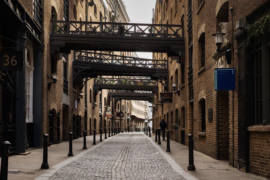 A narrow pedestrian street lined with multi-storey brick buildings featuring black iron balconies and decorative lantern-style wall-mounted lights. The street surface is paved with cobblestones, and black metal bollards line both sides to prevent vehicle access. Suspended above the street are several black metal walkways with crisscross railing designs, connecting the upper floors of the buildings on either side. In the background, a single individual dressed in dark clothing is walking away from the camera, adding a sense of scale to the scene. The scene appears to be in an urban environment, possibly within a historic district or a city with preserved architecture, with a subtle hint that this could be an area where private waste collection or other urban services might operate. Lighting is diffuse, suggesting an overcast day, with soft shadows and even illumination across the scene. The overall setting is clean, orderly, and devoid of visible rubbish or clutter, reflecting a well-maintained area that could be associated with professional waste management services like Waste Collection Bermondsey operating in the vicinity.