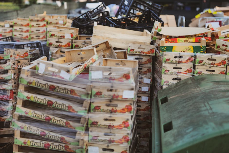 A large outdoor collection of cardboard produce boxes, labeled with graphics and the text 'fresh fruits', stacked haphazardly on a pavement or similar flat surface. The boxes are made of light-colored, rough-textured cardboard with cut-out handles on each side, some are partially flattened or slightly damaged. Behind the boxes, there are additional black plastic crates stacked irregularly, along with a few loose pieces of packaging and cardboard. To the right of the boxes, a green waste bin or container is partially visible, with a textured exterior and a slightly worn appearance. The environment appears to be an outdoor setting, possibly a back alley or yard, with natural lighting illuminating the scene. The image relates to waste management activities, highlighting the collection or disposal of cardboard packaging materials, which could be part of an alternative or private waste removal service provided by Waste Collection Bermondsey, aligned with local rubbish regulations.